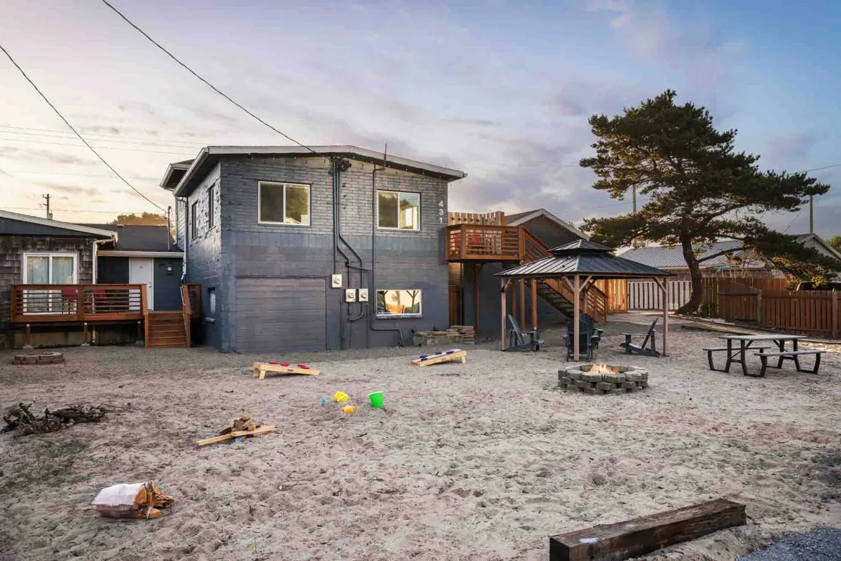 This image captures the inviting exterior of a two-story cottage set in a sand-lined yard. Prominently featured are a cozy fire pit, a picnic table perfect for outdoor dining, and a play area scattered with toys. The scene is lively yet relaxed, suggesting evenings spent around the fire or casual gatherings under open skies.
