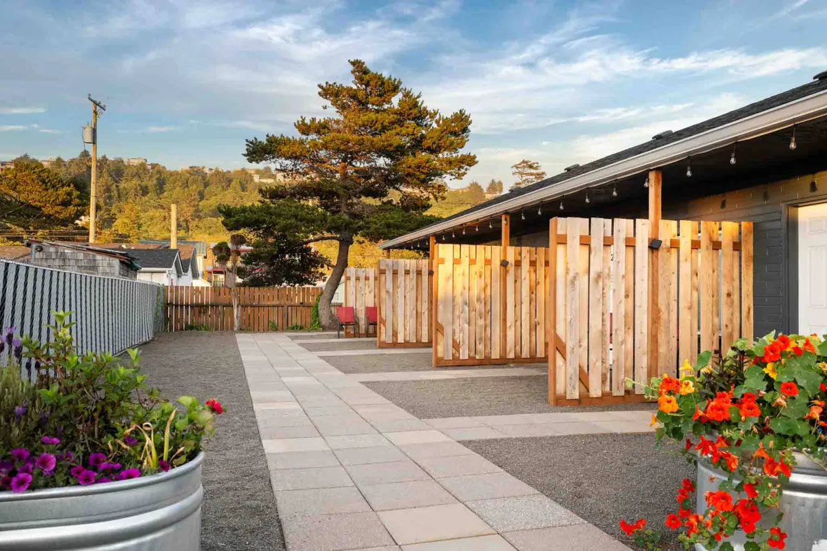 An inviting outdoor common area featuring wooden fences, potted flowers, and a pathway. The scene suggests places for guests and their pets to stroll and relax, with natural greenery and outdoor seating options under the trees.