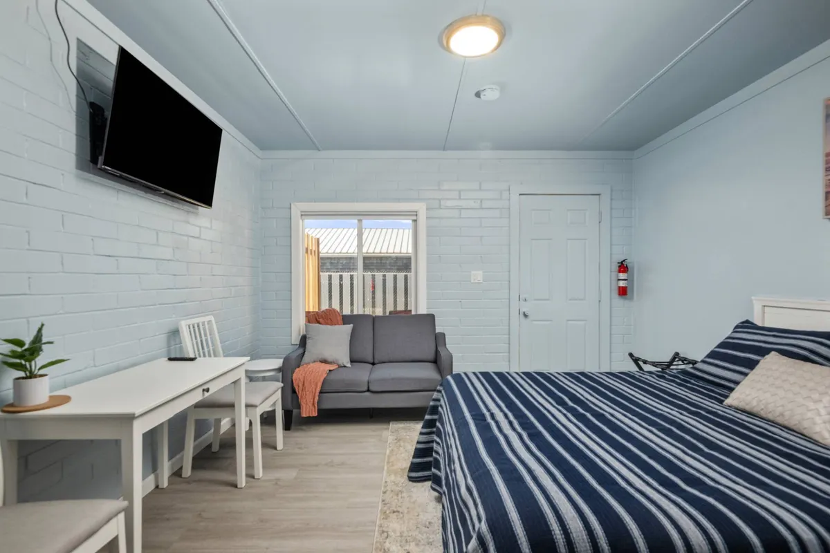 A cozy cottage interior with a striped bedspread, sofa, wall-mounted TV, window bringing in natural light, and a small table with chairs. The scene exudes warmth and comfort, inviting both people and pets to relax and unwind together after a day of adventures.