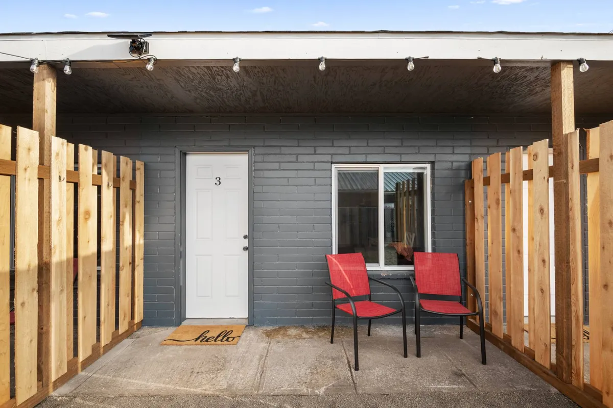 This image captures the entrance to a cottage patio with red chairs, a welcoming 'hello' doormat, and a wooden fence. The setting feels intimate and friendly, suggesting a space perfect for morning coffee or casual chats with neighbors.