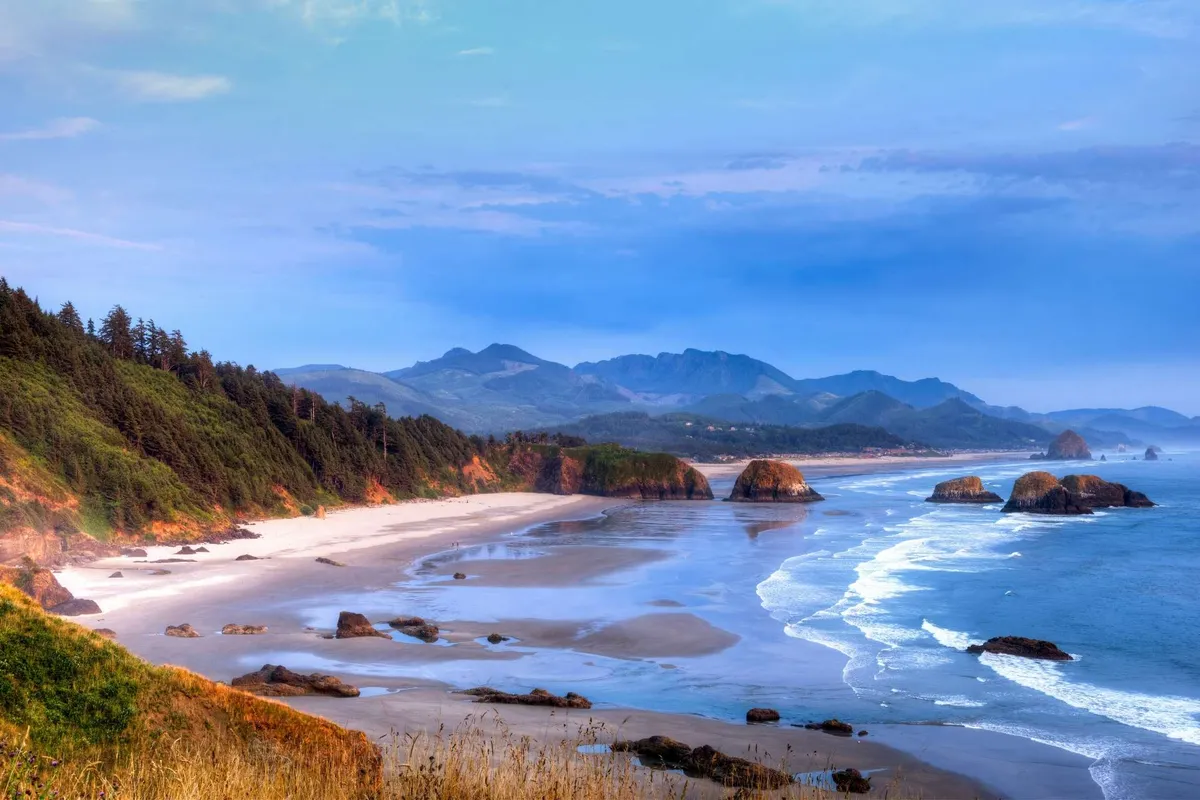 A sweeping view of the Oregon Coast, showcasing gentle waves lapping against a sandy beach, dramatic rock formations, and distant mountains under an expansive sky. The tranquil, natural beauty encapsulates the quintessential coastal experience.