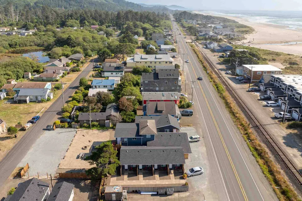 A wide exterior shot showing easy access to the beach, coastal homes, a main road, lush greenery, and a nearby parking area. The scene conveys the seamless connection between the cottages and the surrounding natural landscape—a perfect setup for walks and exploration with a pet.