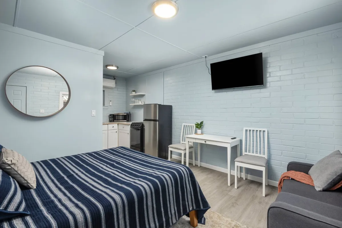 The image showcases the interior of a recently renovated condo suite, featuring a cozy bed adorned with a striped blanket, a quaint dining table with chairs, a modern kitchenette, a decorative mirror, and a TV mounted on the wall. The space exudes warmth and comfort, inviting guests to relax, enjoy a homemade meal, or unwind with their favorite show.