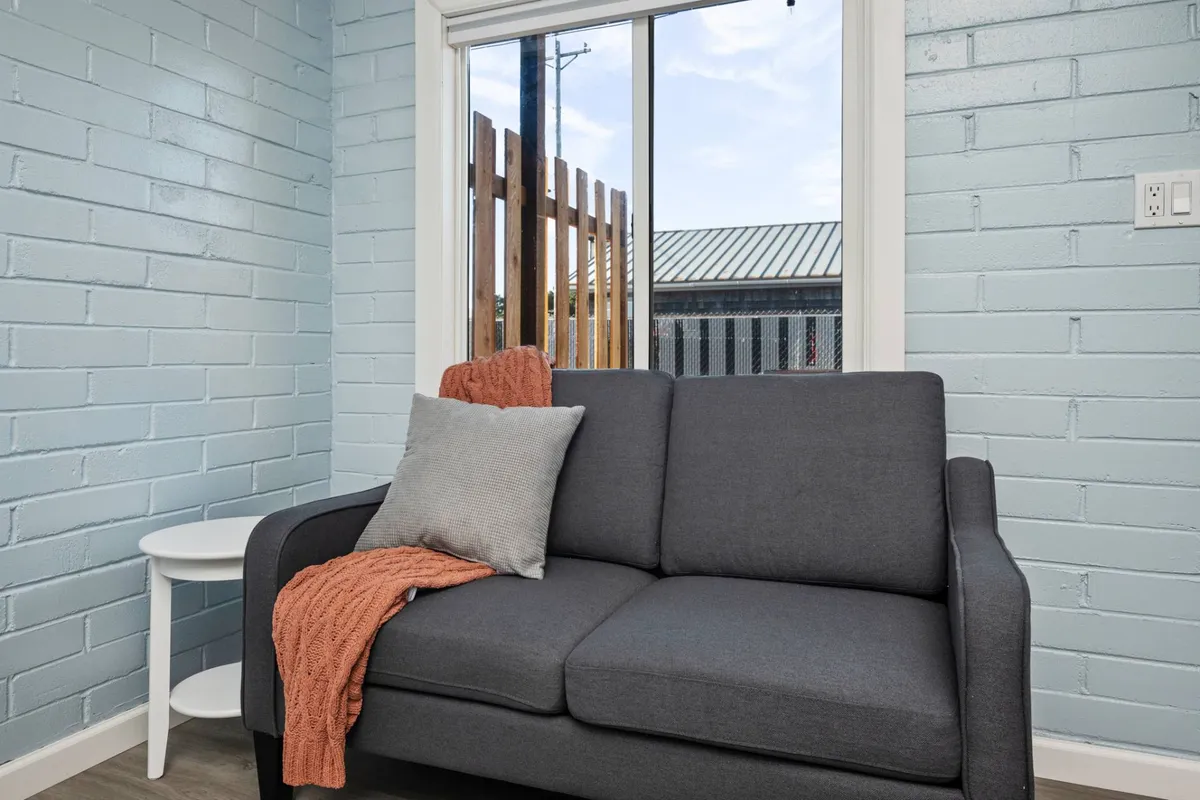 A comfortable living area featuring a gray sofa with an orange throw blanket and decorative pillow, positioned by a window that lets in ample daylight. The setting is perfect for relaxing or enjoying a quiet moment.