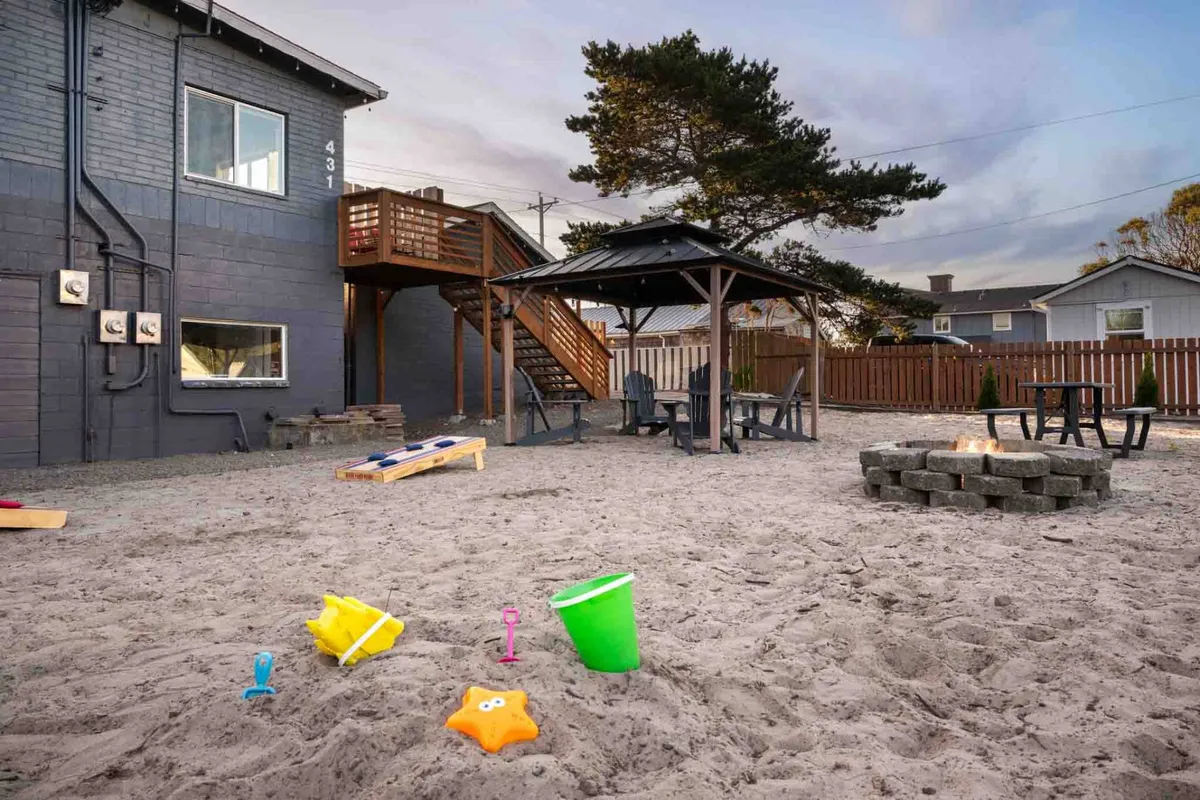 A lively outdoor sand area featuring toys, a covered seating space (gazebo), a fire pit, and a staircase to the upper level. The scene captures a communal and playful environment perfect for families and pet owners alike.